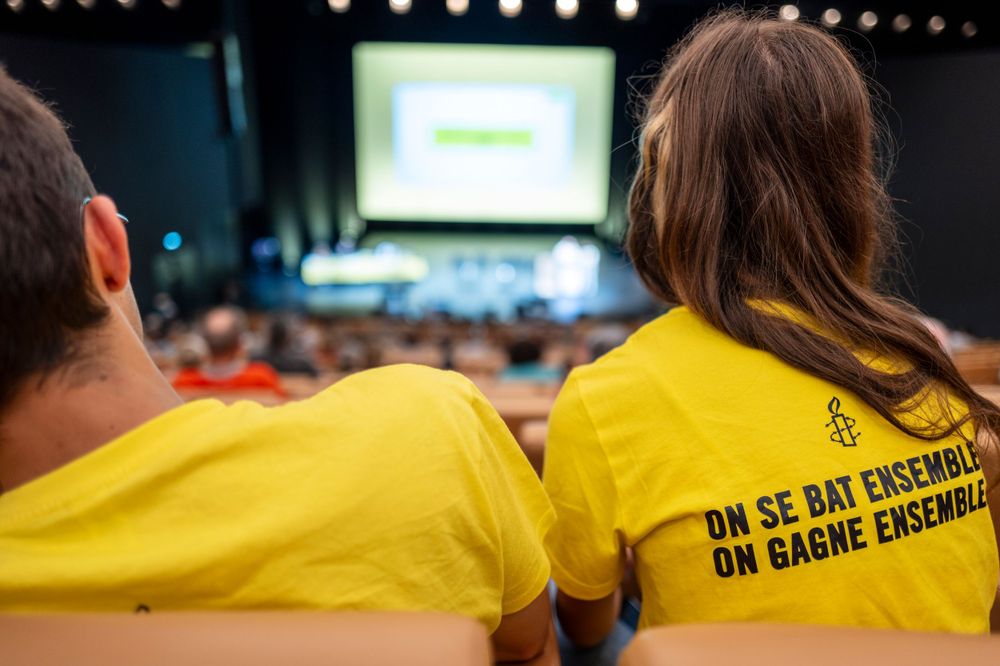Two people wearing yellow t-shirts with French text sit in an auditorium facing a presentation screen.
