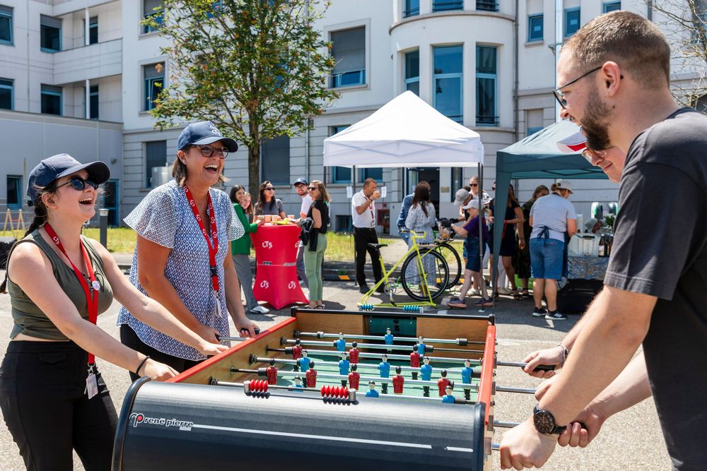 People gathered outdoors playing foosball tables at a social event with white tents and buildings in the background.