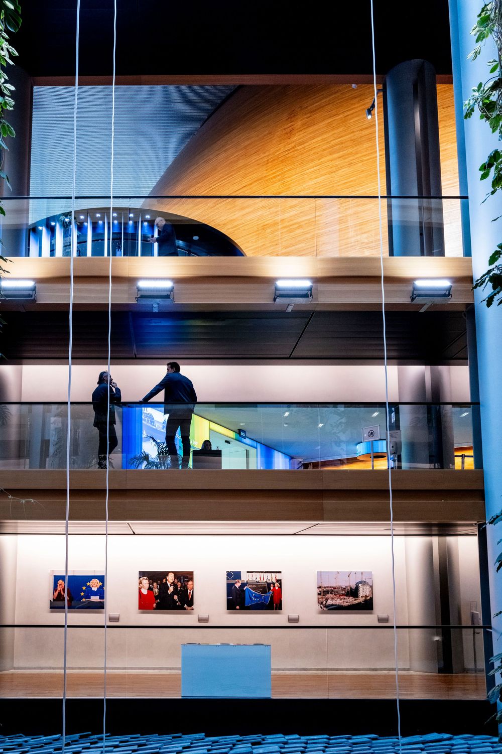 Modern architectural interior with curved wooden ceiling, glass walls and silhouetted figures on upper level viewing art gallery.