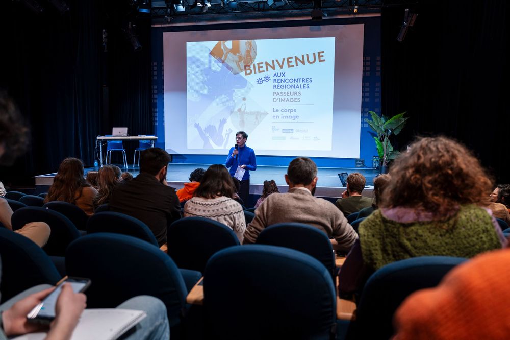People seated in a dark auditorium watching a presentation with a welcome message displayed on a large screen.