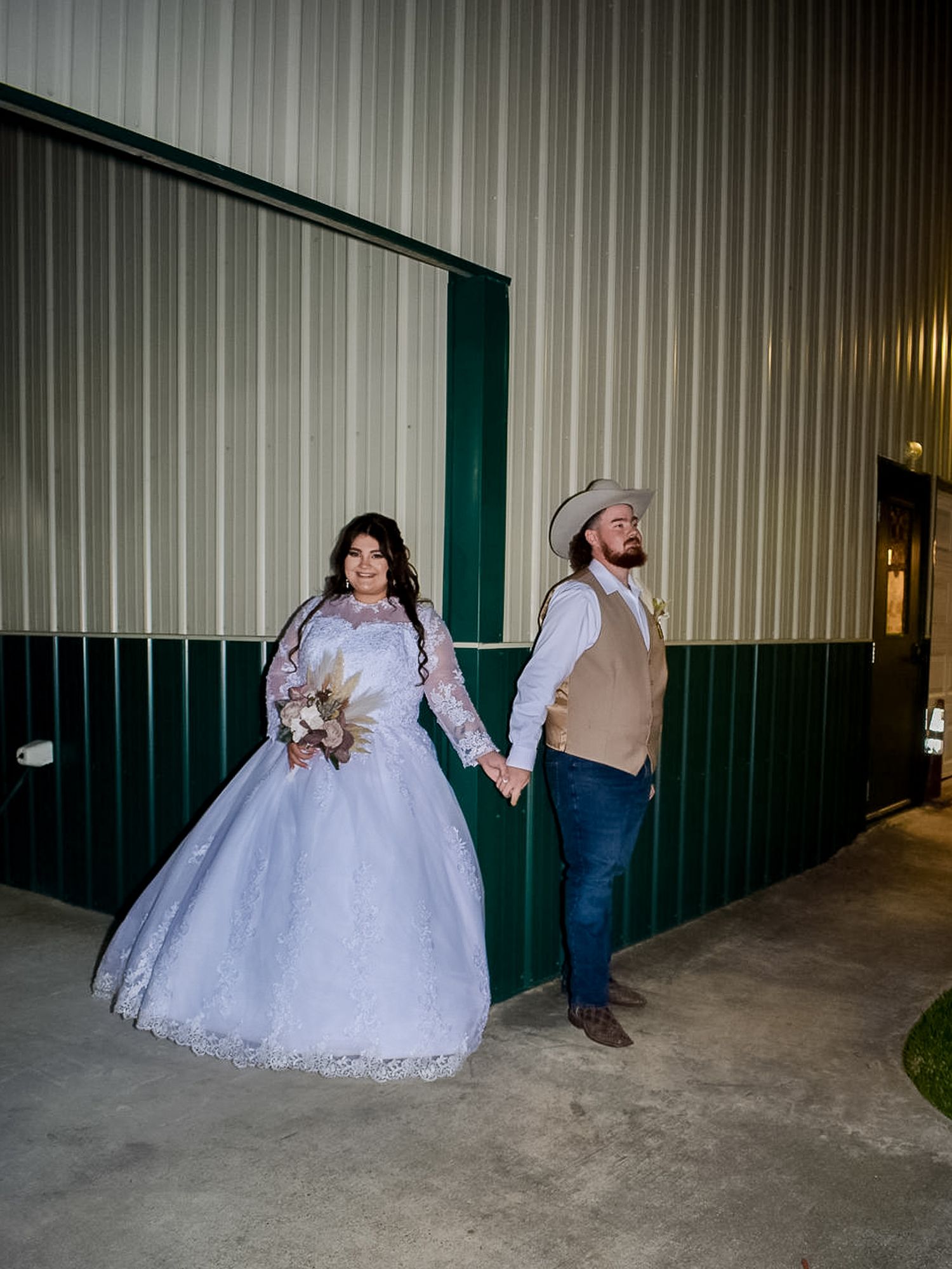 A couple in wedding attire holding hands against a metal wall with white dress and casual groom style.