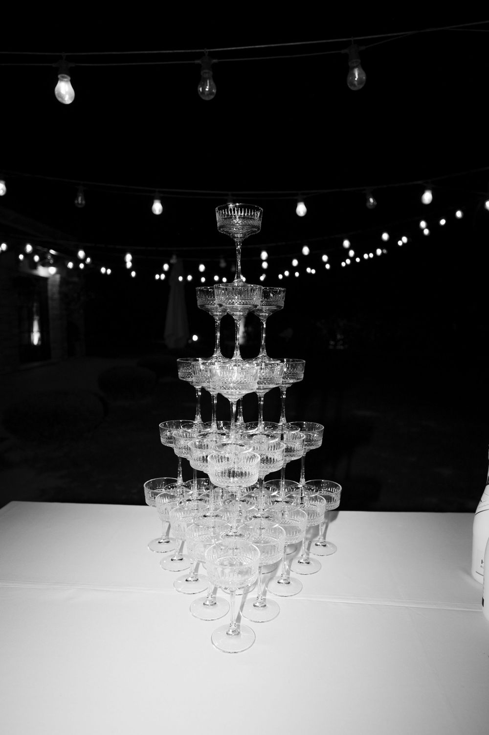 A champagne tower with multiple tiers of crystal glasses illuminated by overhead string lights in a dark setting.