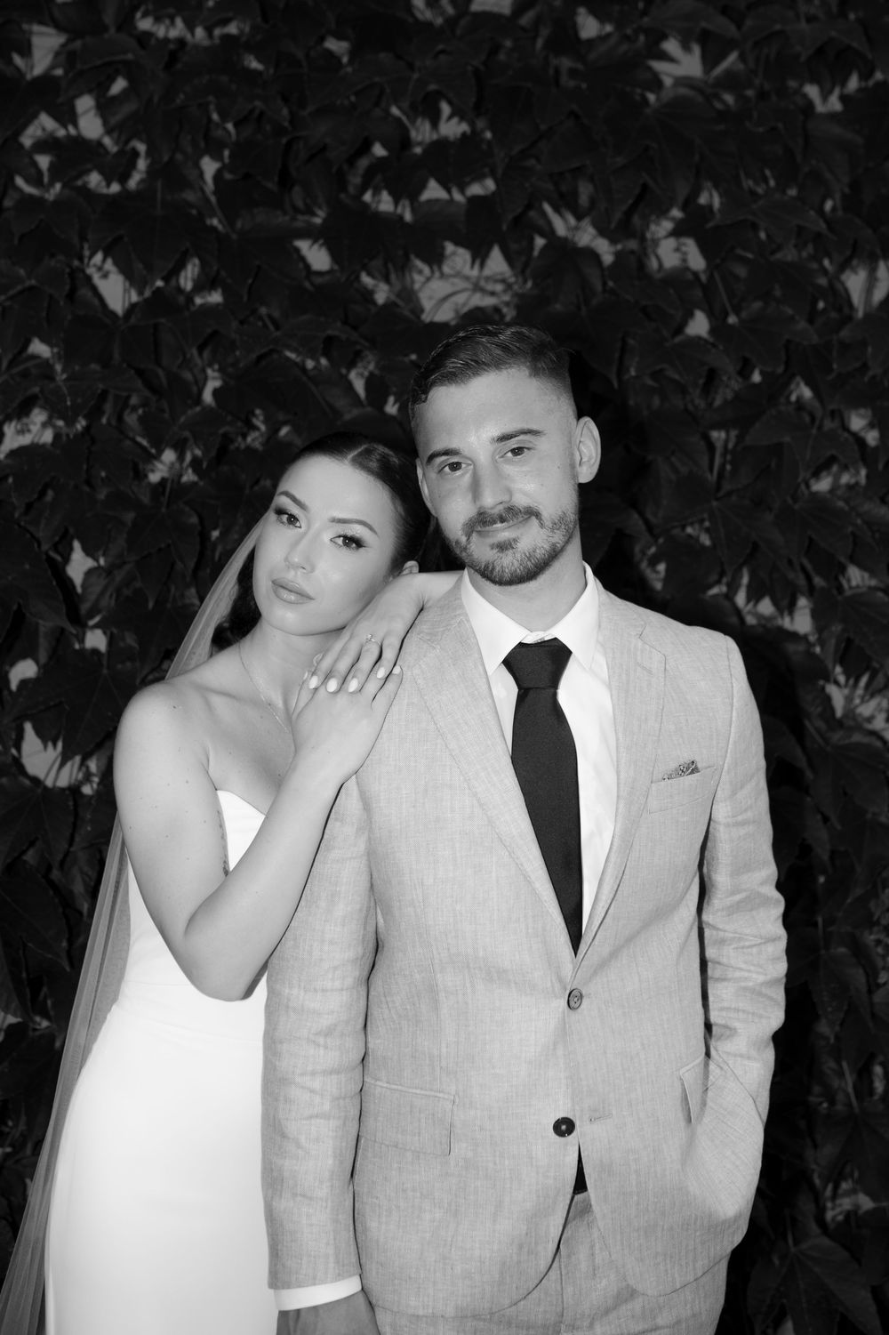 A couple poses for romantic black and white wedding portraits against a leafy backdrop.