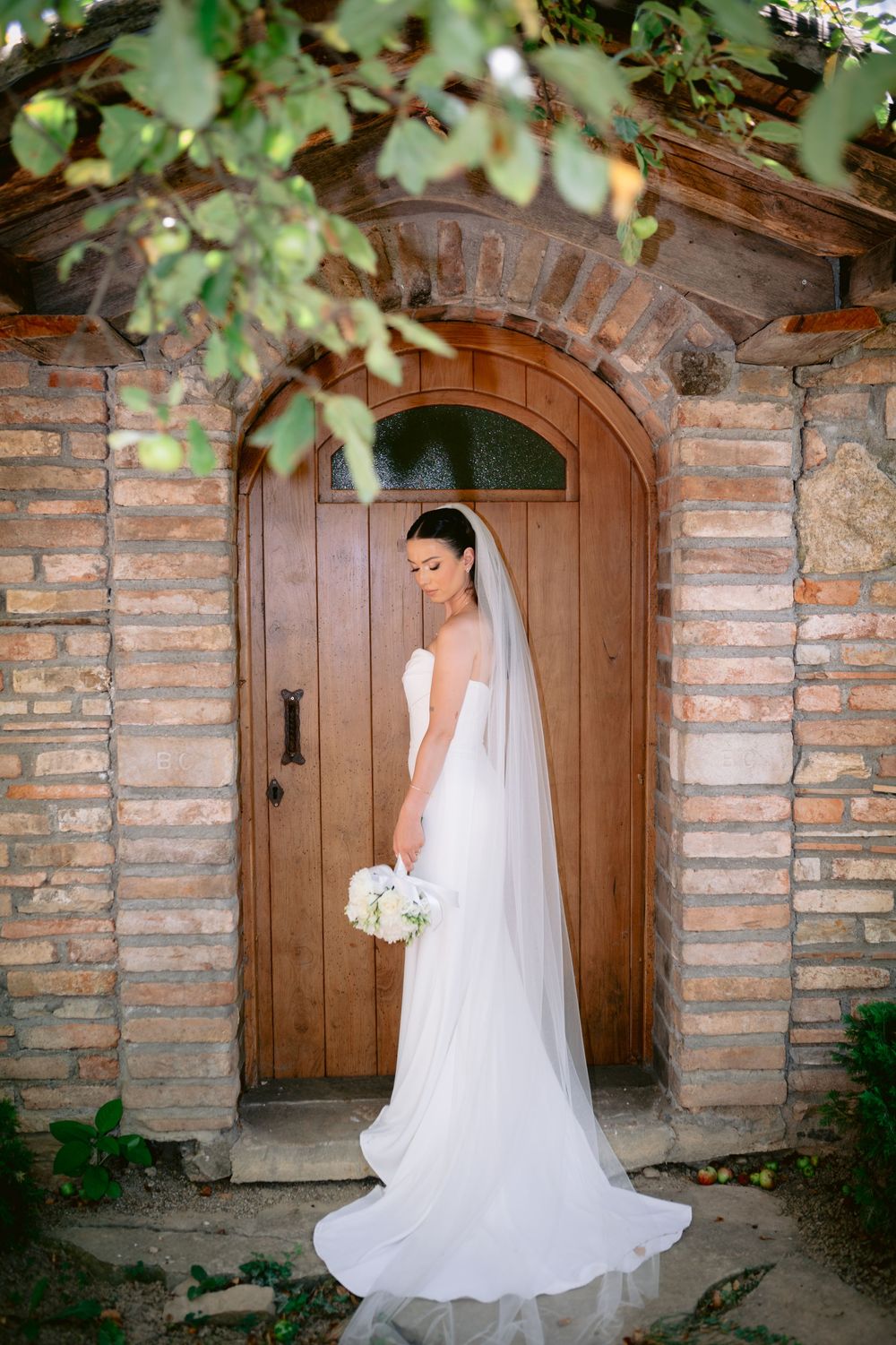 Long white wedding gown and veil flow elegantly in archway of rustic stone and brick doorway surrounded by greenery.