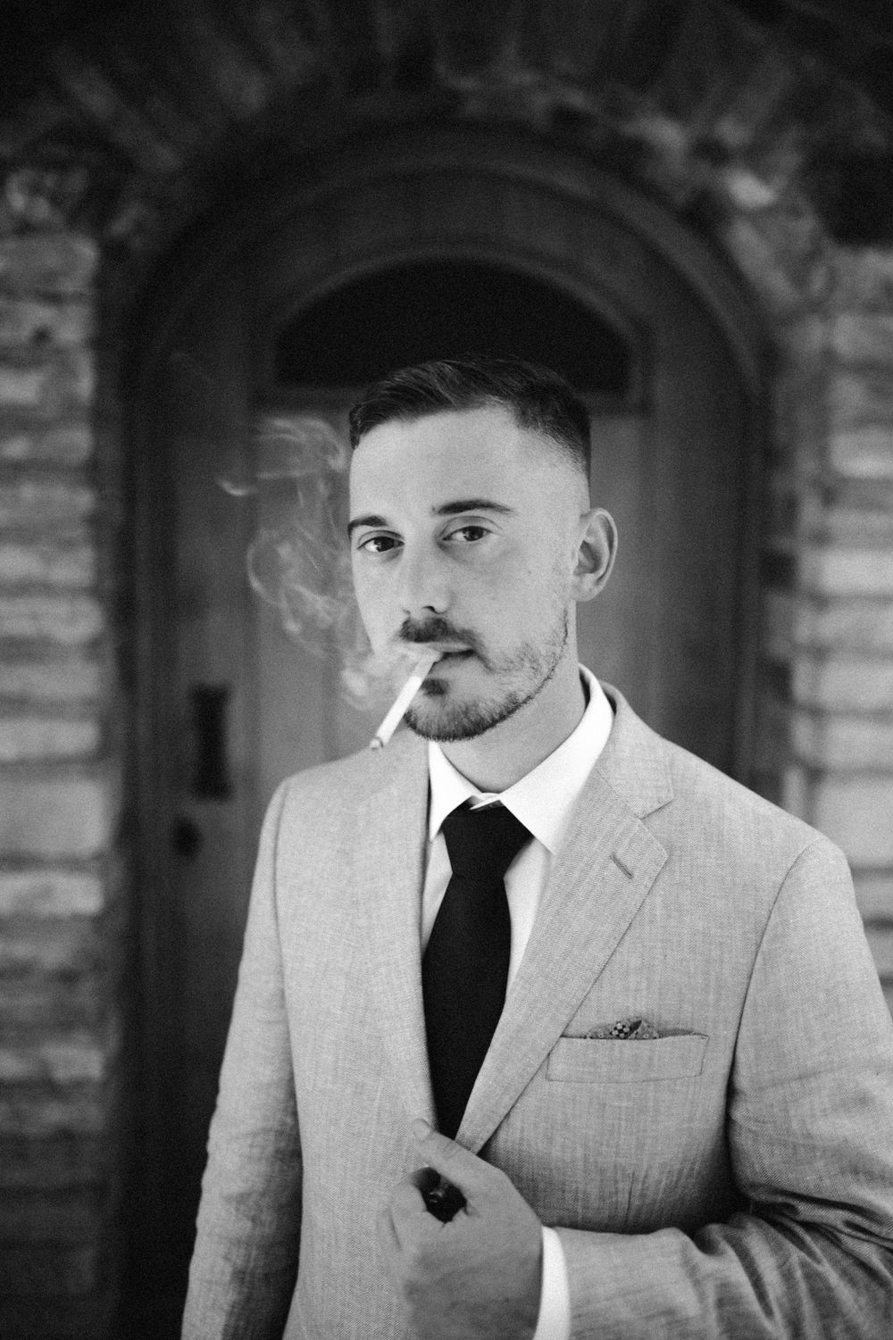 Black and white portrait series of a groom in a light suit and black tie smoking.