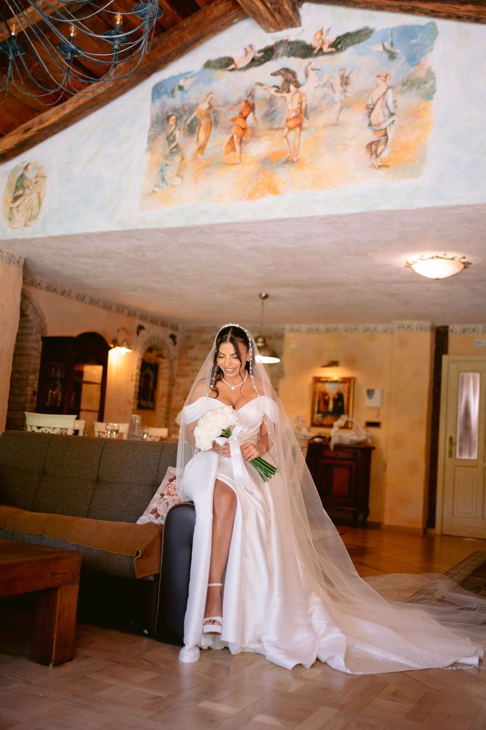A beautiful bride in a flowing white gown and veil poses against a wall with painted murals in an elegant indoor setting.