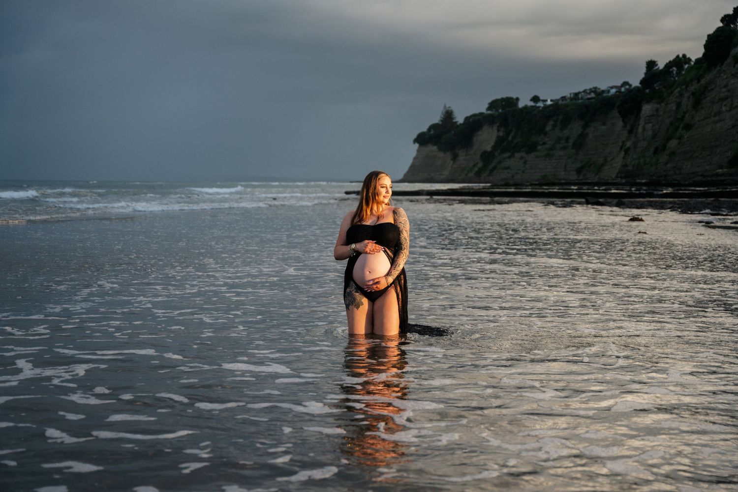 A silhouette standing in shallow ocean water against a cloudy coastal backdrop.