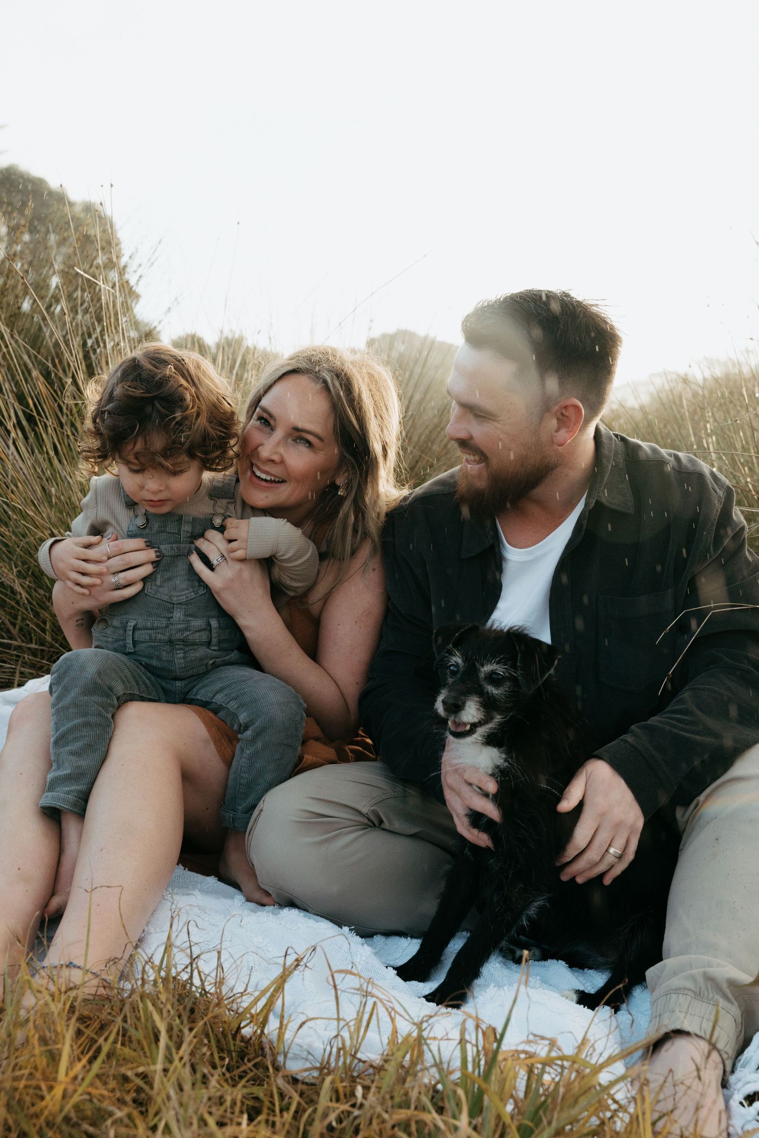 A family sits together on a blanket outdoors with their black dog during sunset.