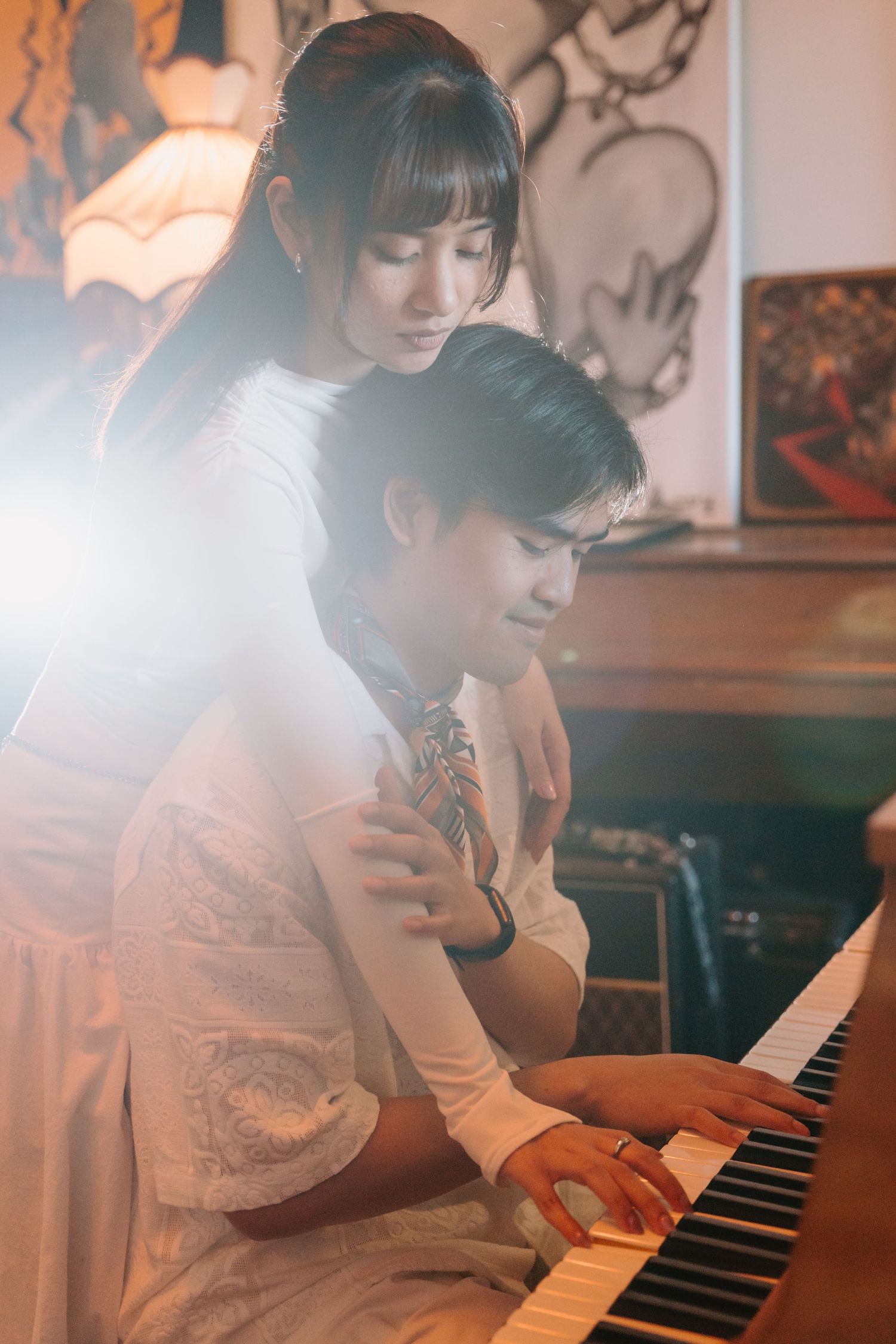 Two people share an intimate moment at the piano in a softly lit room with vintage decor.
