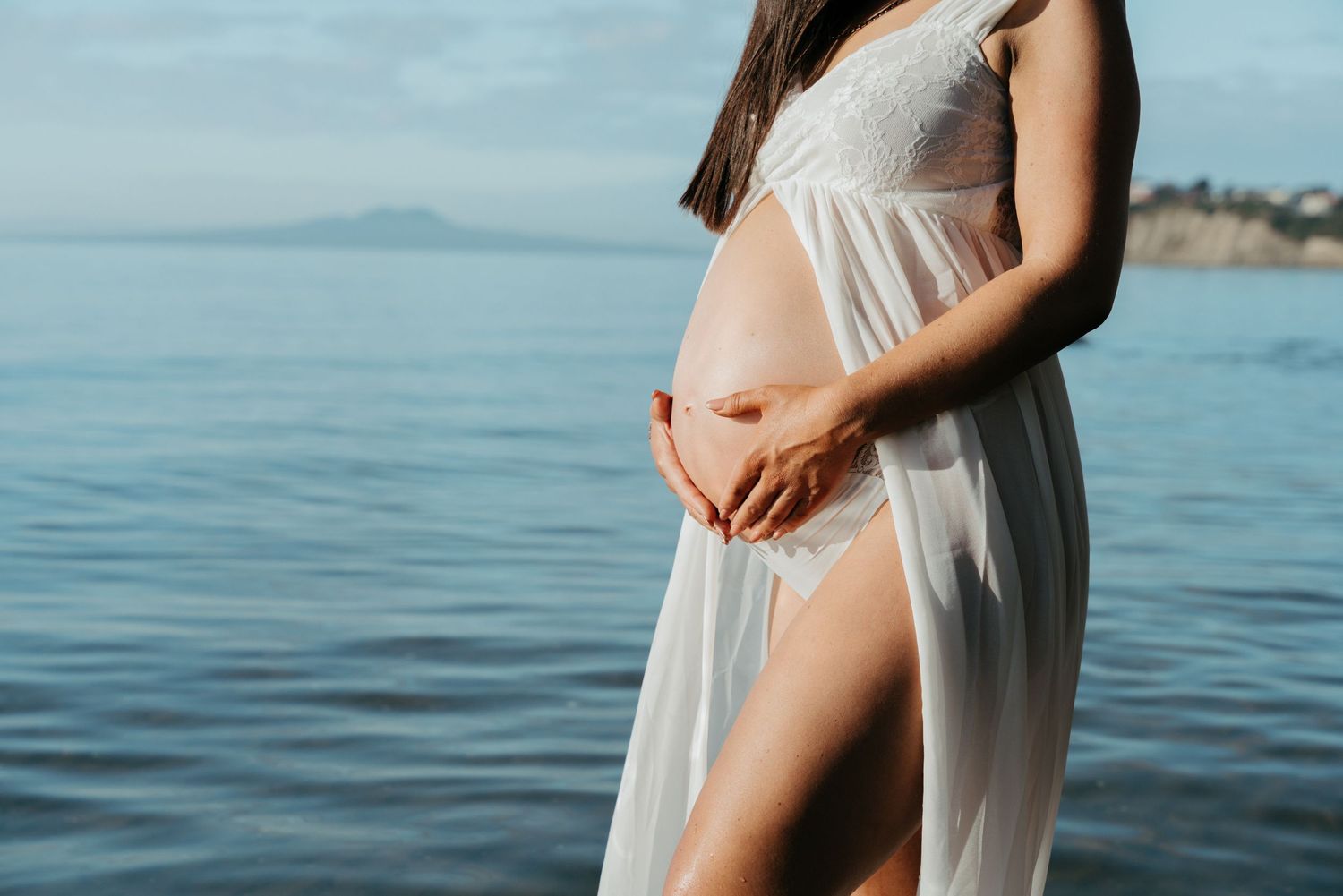 Silhouette of a pregnant figure in a flowing white dress standing by the ocean at sunset.