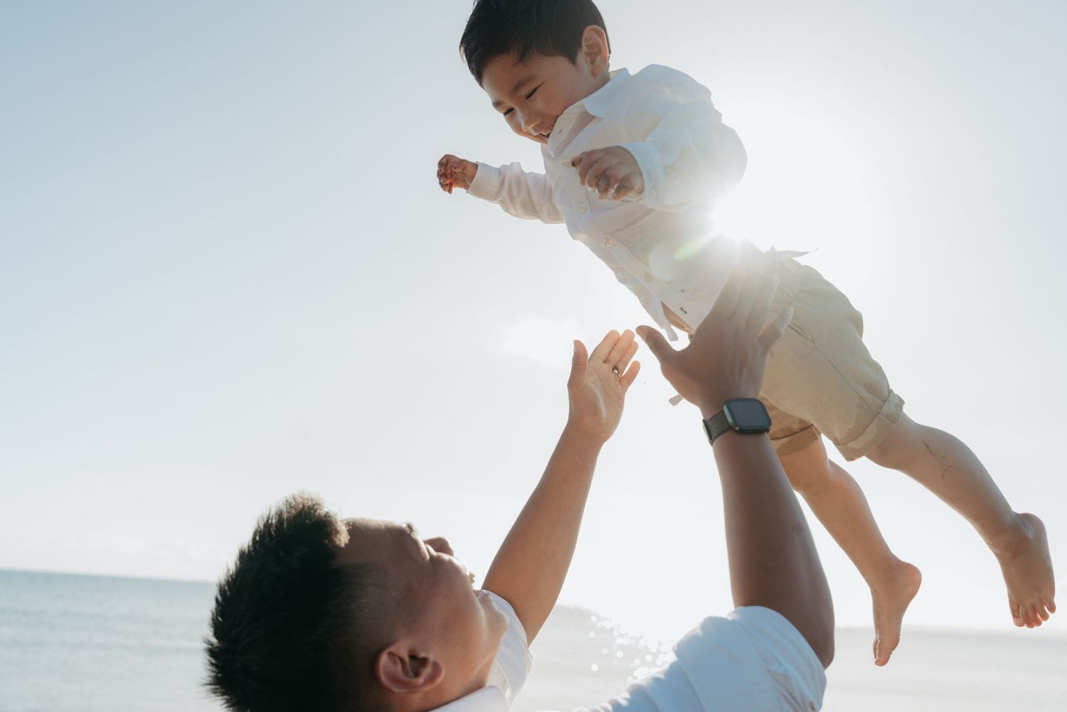 Parent lifting child high into the air against bright sunlight at the beach.