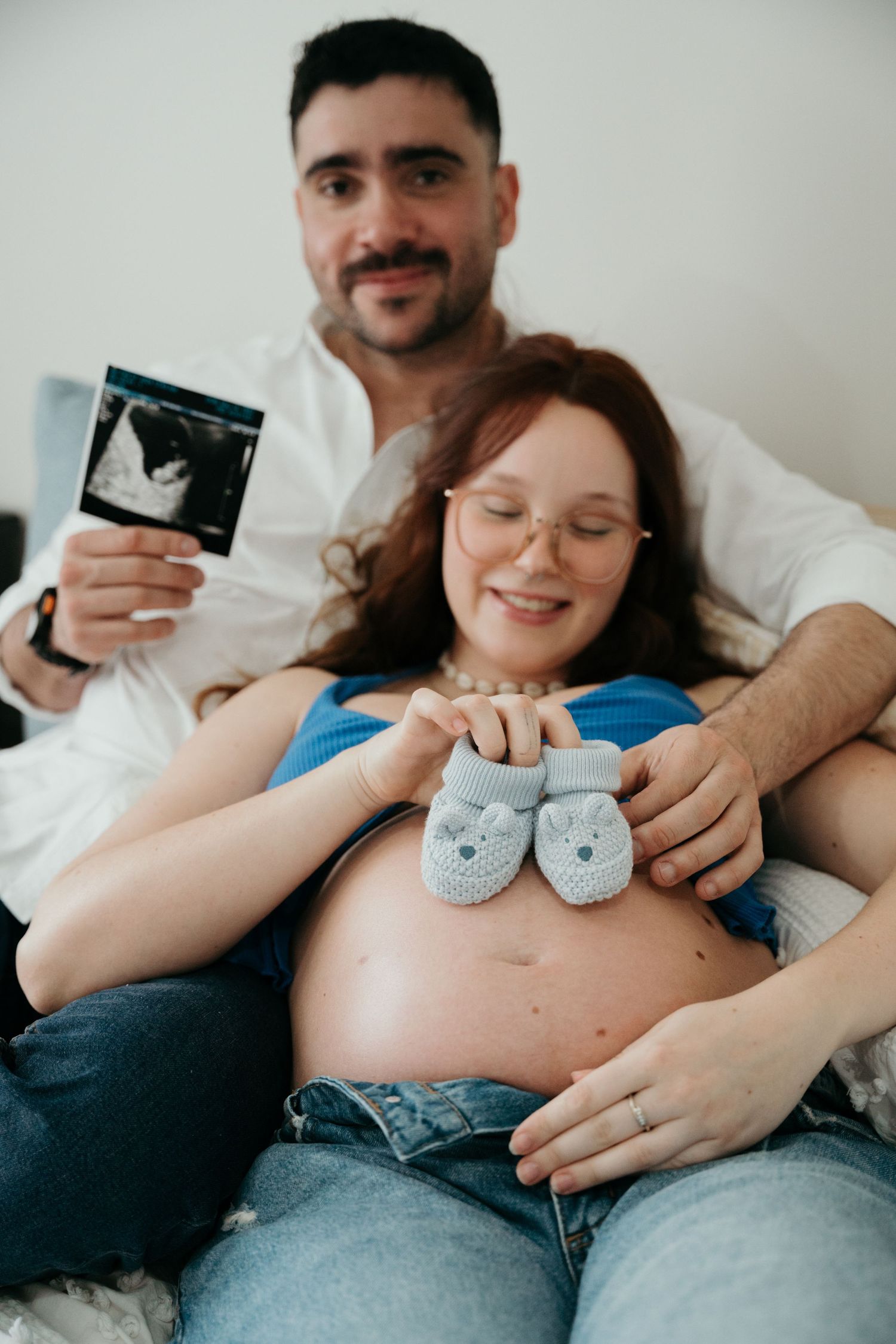 Expectant couple holding baby shoes and ultrasound photo while relaxing on bed.