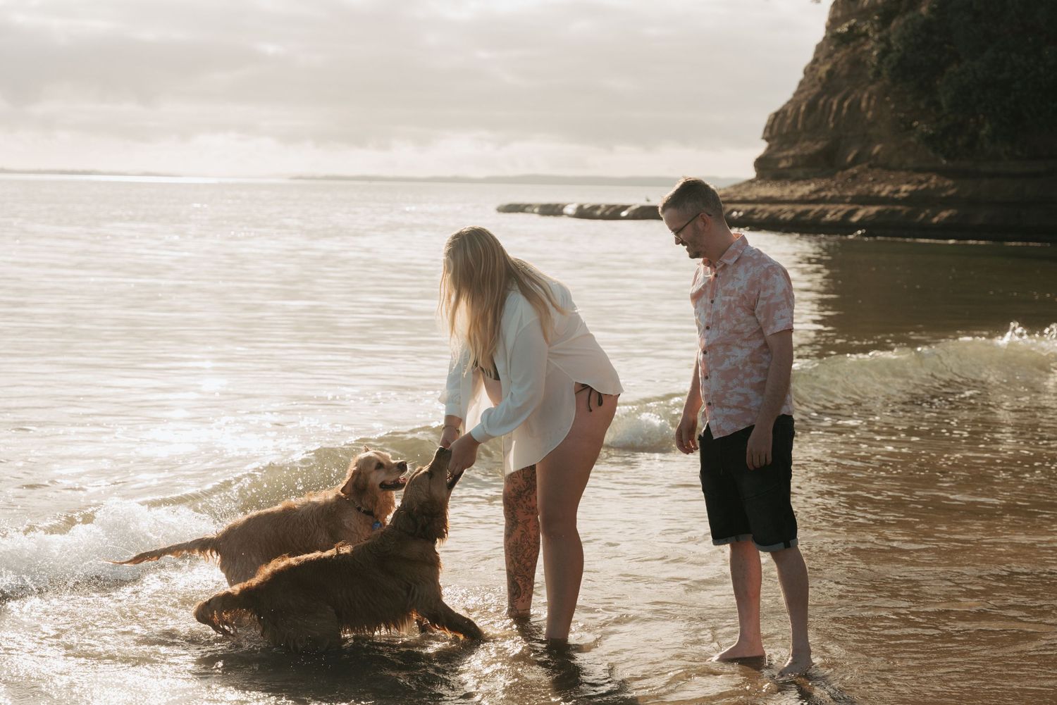 A couple plays with their dog on a misty beach at sunset near rocky cliffs.