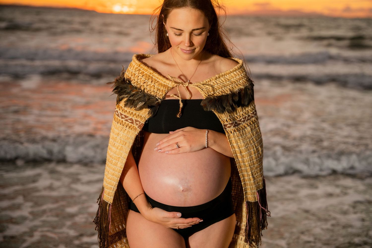 An expectant mother in a knit cardigan poses on beach at sunset.
