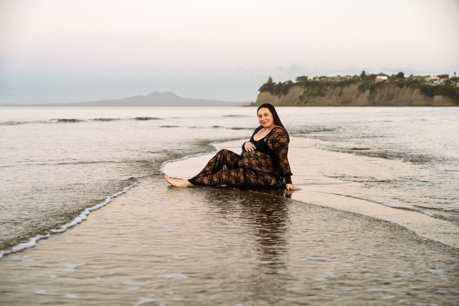 A woman in flowing dress sits in gentle ocean waves during sunset.