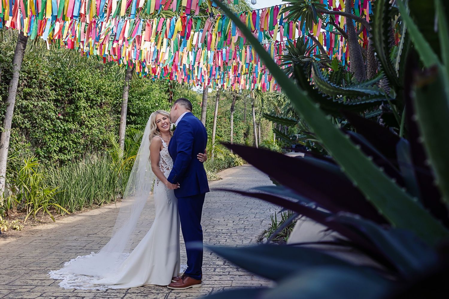 A couple poses for their wedding photos under vibrant prayer flags in a garden pathway.