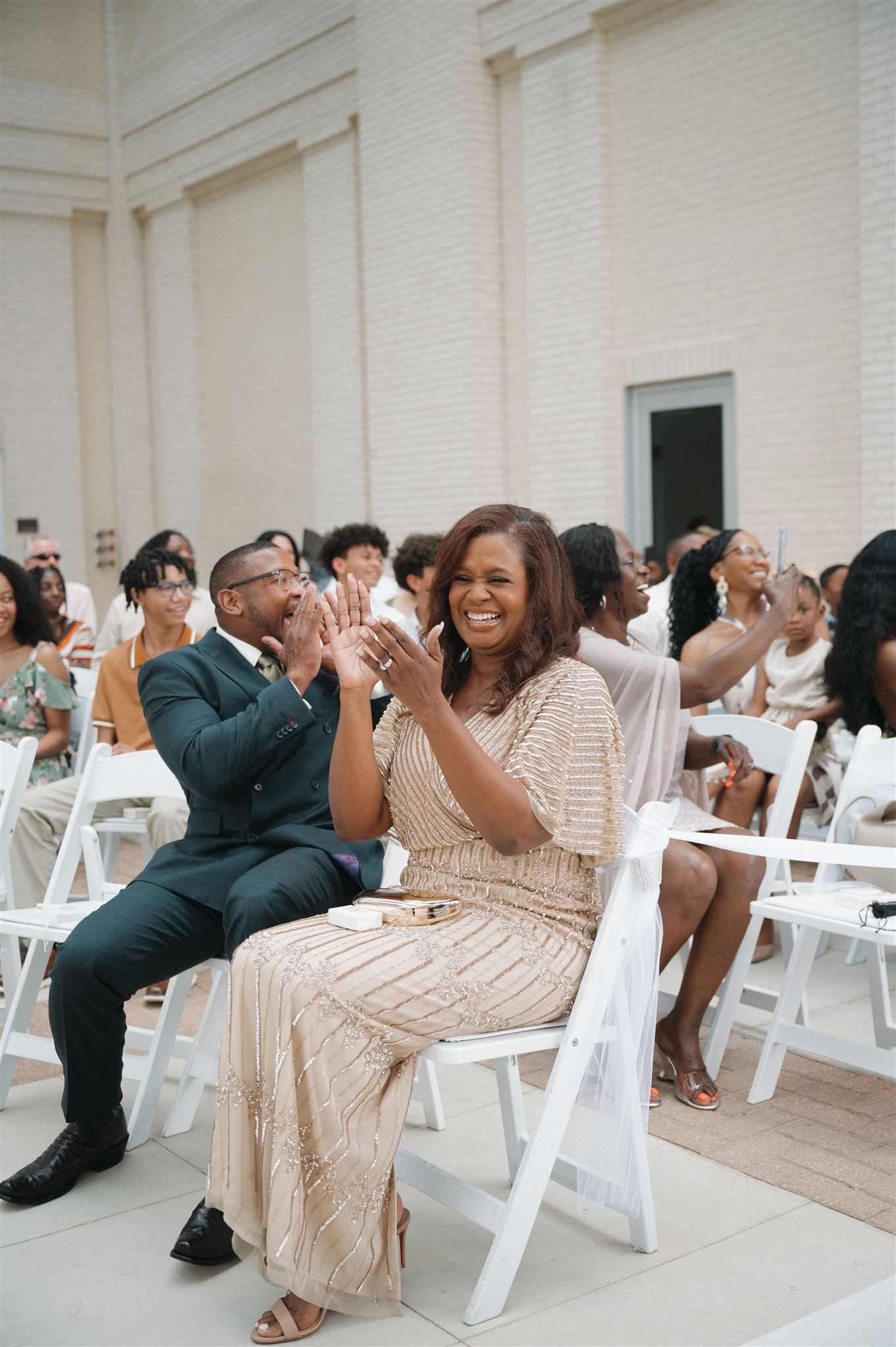 Wedding guests sitting in white chairs clap and laugh together during an outdoor ceremony.