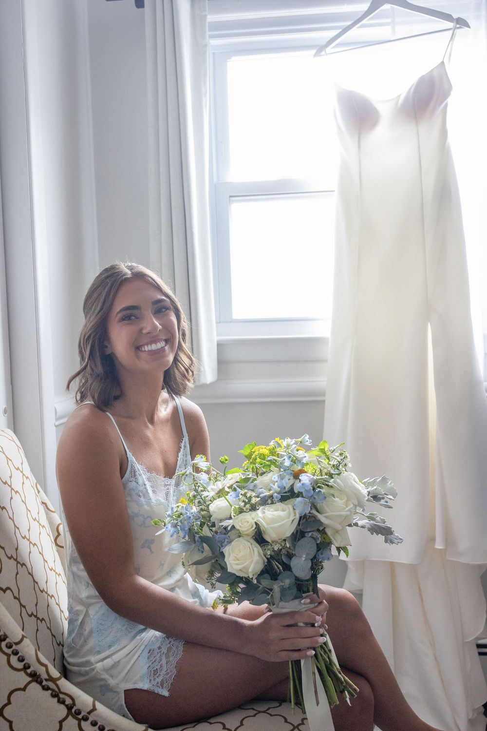 Smiling bride in lingerie holds white and blue bridal bouquet beside hanging wedding dress near window.