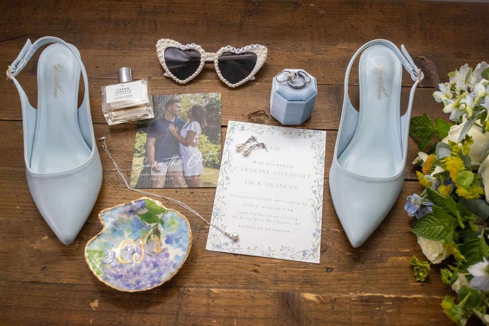 Flat lay of wedding details including blue heels, heart sunglasses, invitation, rings, and bridal bouquet.