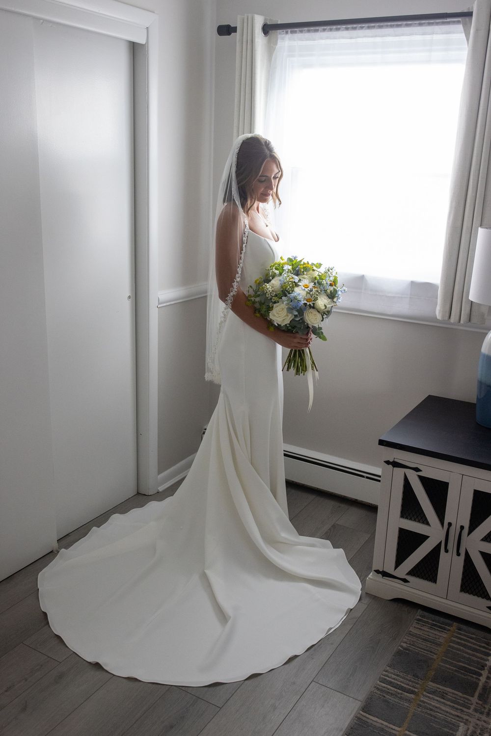 Elegant bride in backless white gown with cathedral veil holds bouquet while standing by bedroom window.