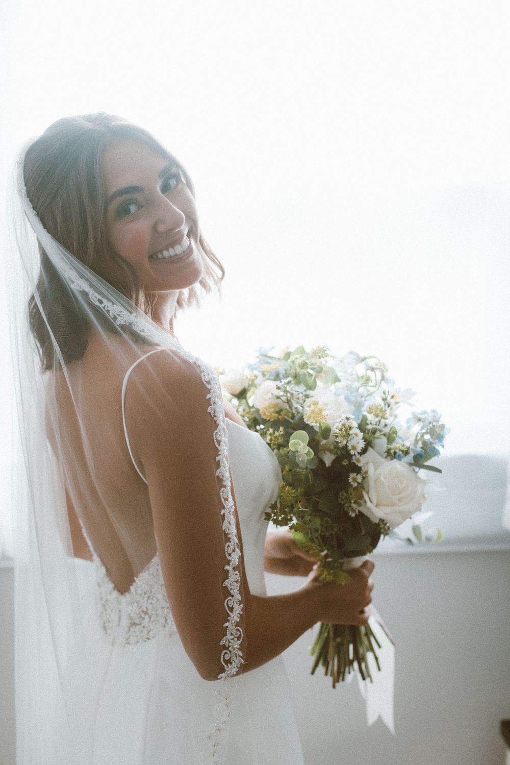 Radiant bride in lace-back wedding gown smiles over shoulder while holding white and blue floral bouquet.