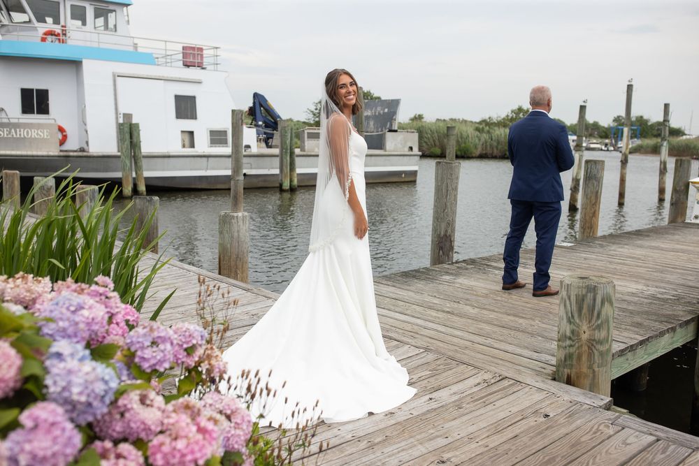 Bride in flowing white gown stands on waterfront dock as groom awaits first look moment ahead.