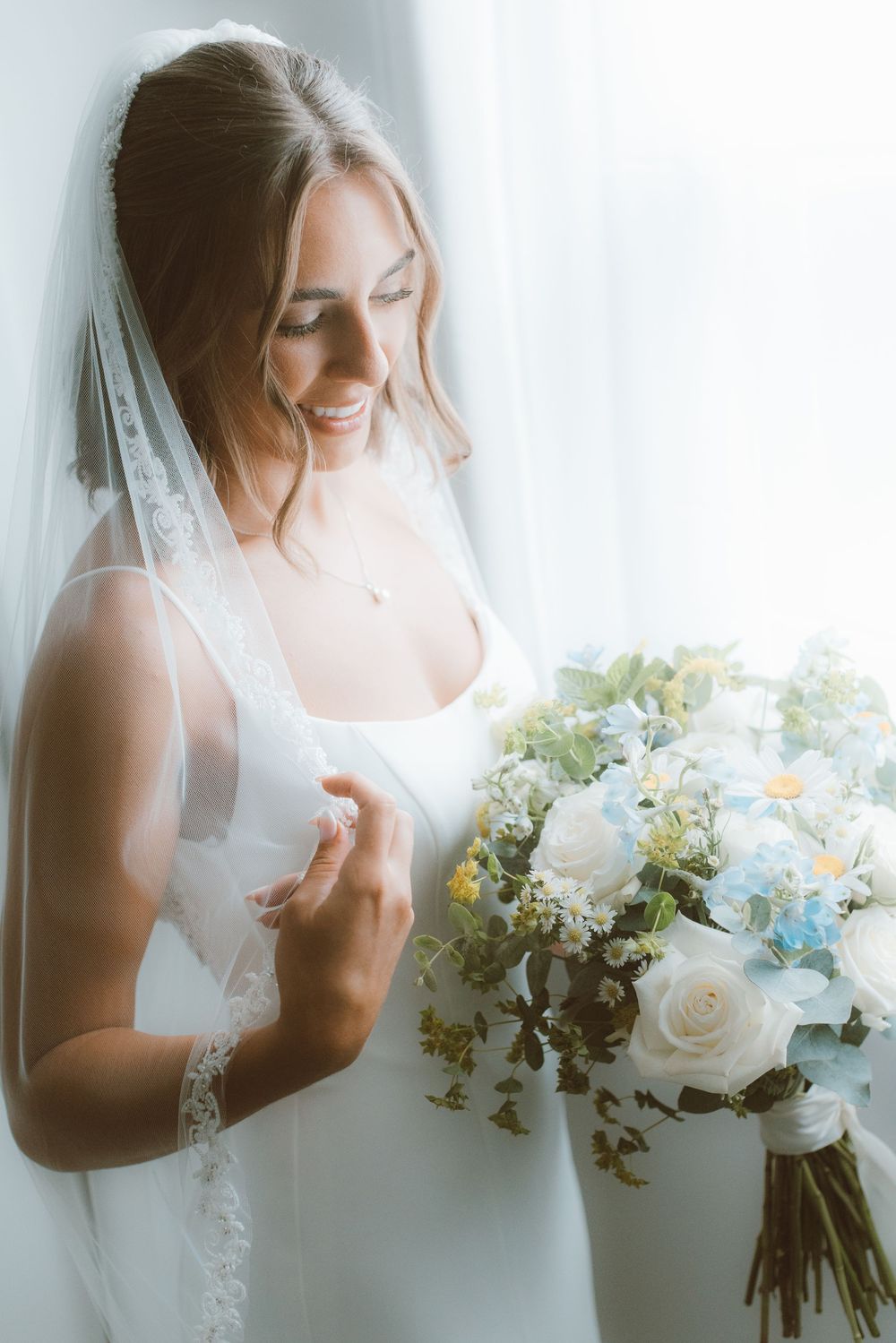 Smiling bride in white gown and lace veil holds a bouquet of white and blue flowers by a bright window.