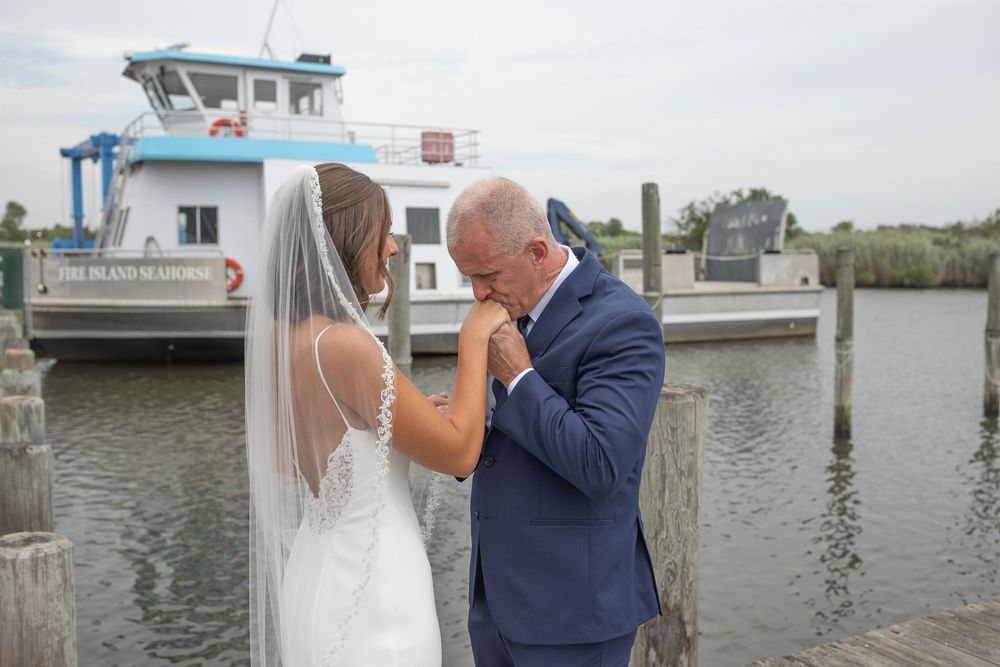 Emotional father kisses brides hand on a dock with a ferry boat in the background.