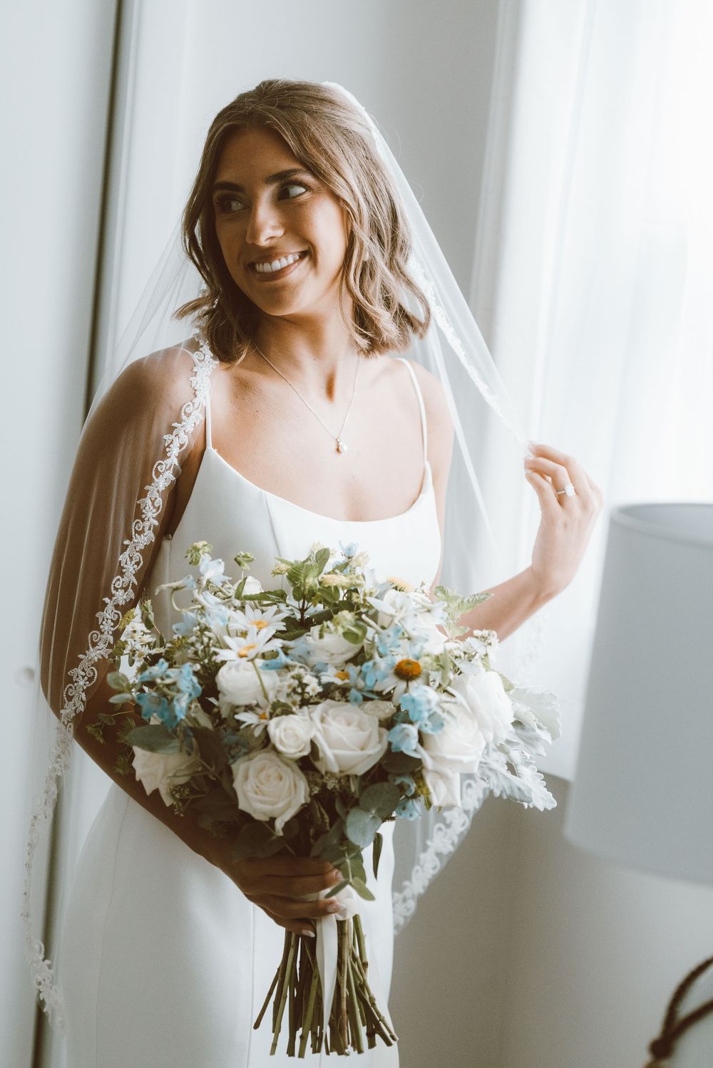Radiant bride in white dress and lace veil smiles while holding a blue and white floral bouquet.