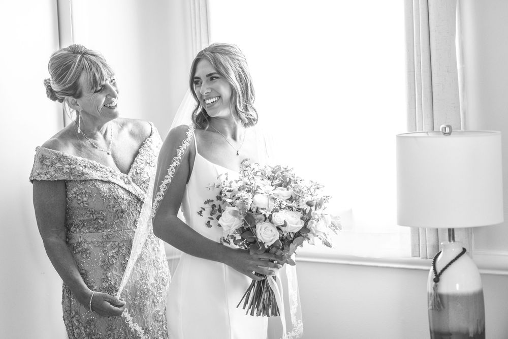 Black and white photo of bride and mother sharing a joyful moment by a window before the wedding.