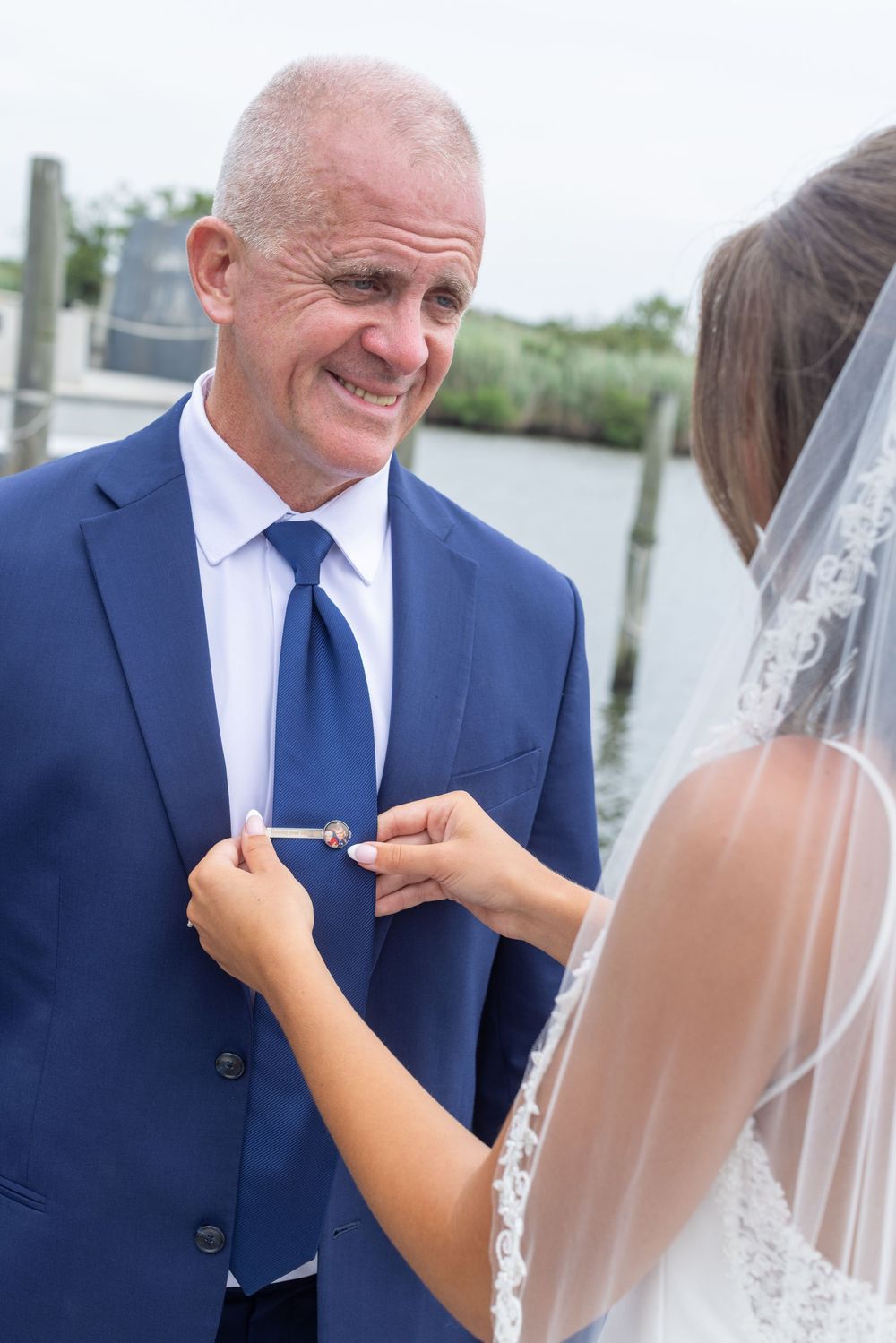 Bride pins a boutonniere on smiling groom in navy suit at a waterfront wedding venue.
