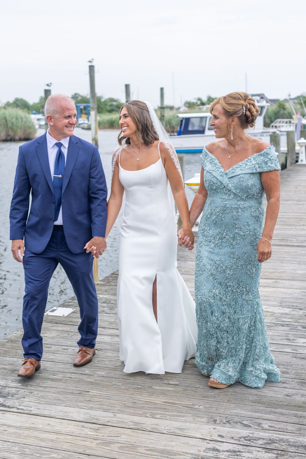 Bride walks hand in hand with parents along a wooden dock at a waterfront wedding.