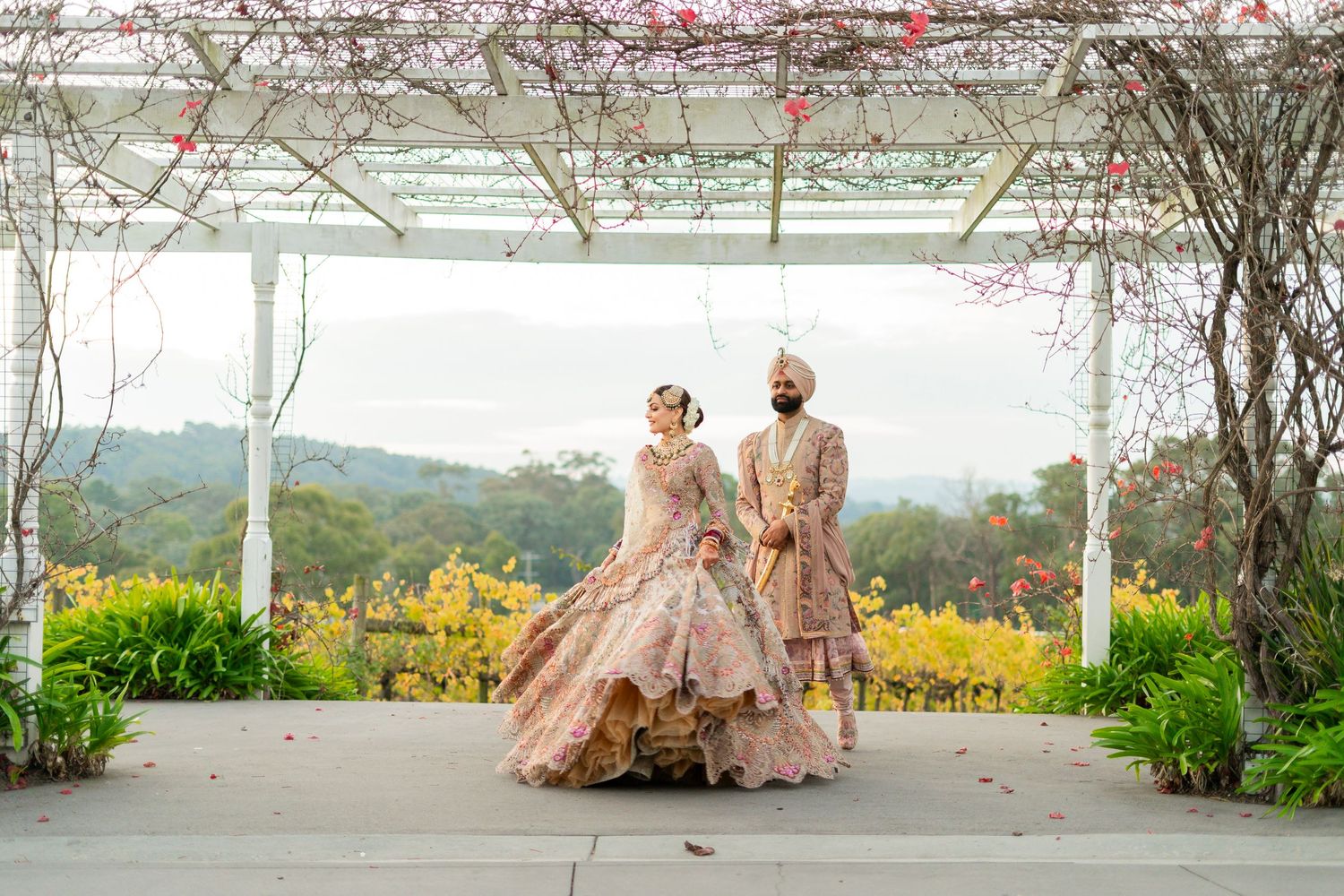 A couple in traditional Indian wedding attire poses under a floral archway with scenic mountain views in the background.
