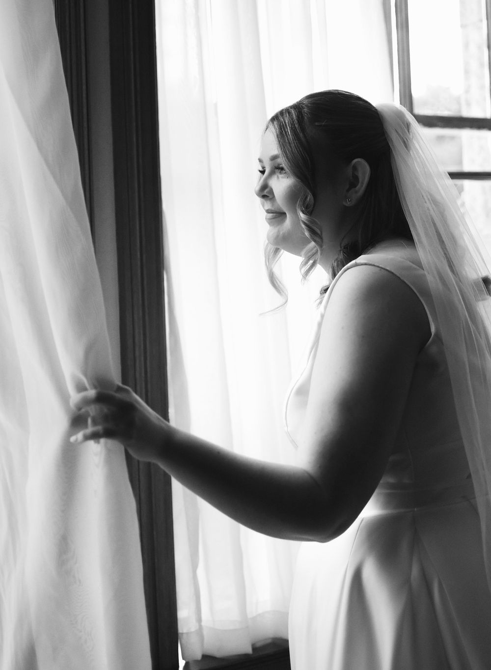 Black and white image of a Bride peeking through the curtains at Balgeddie House Hotel. Natural light against a curtain backdrop.