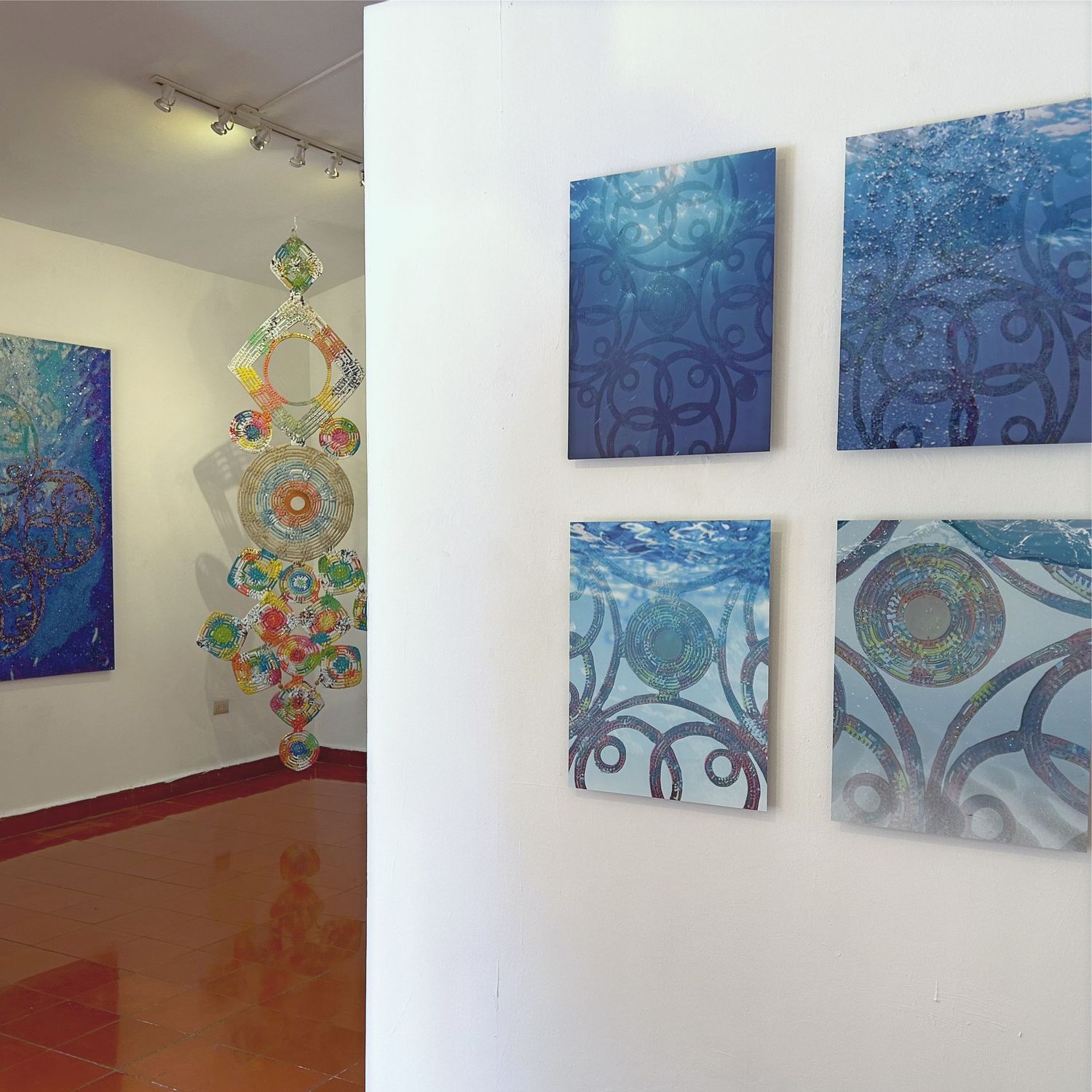 Gallery wall displaying blue abstract paintings and hanging sculptural piece with green elements.