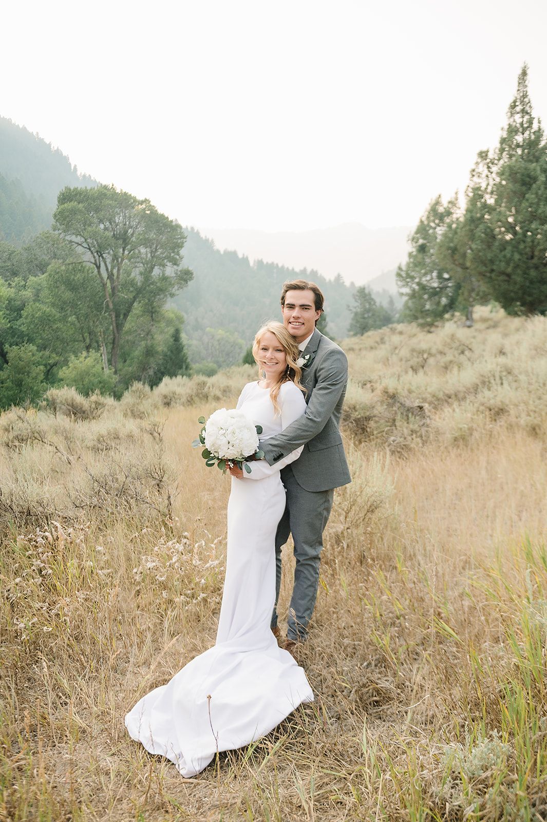 Happy couple embraces in mountain meadow during wedding photoshoot.
