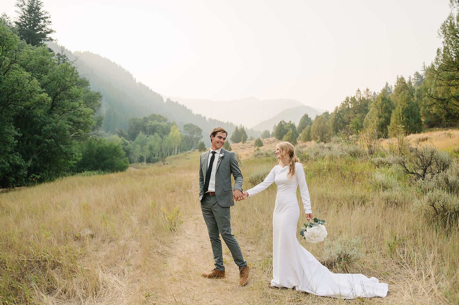 Elegant wedding couple walking together through tall grass in a scenic mountain valley.