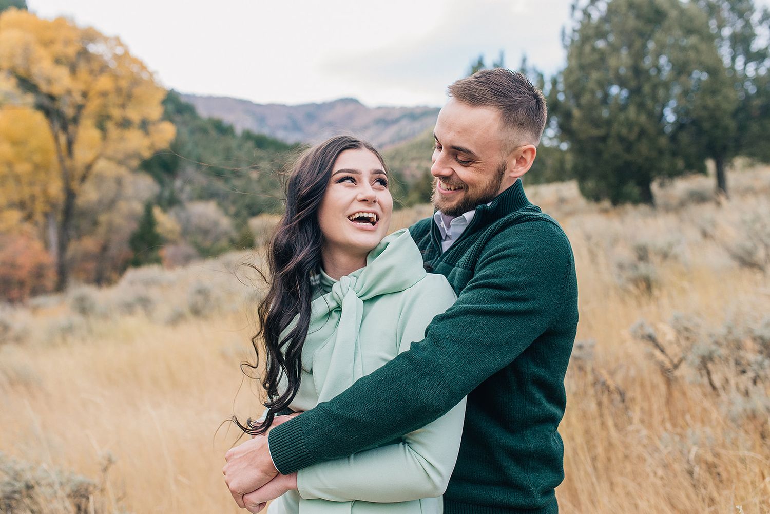 A happy couple embraces and laughs together in an autumn field with golden grass and mountain scenery.