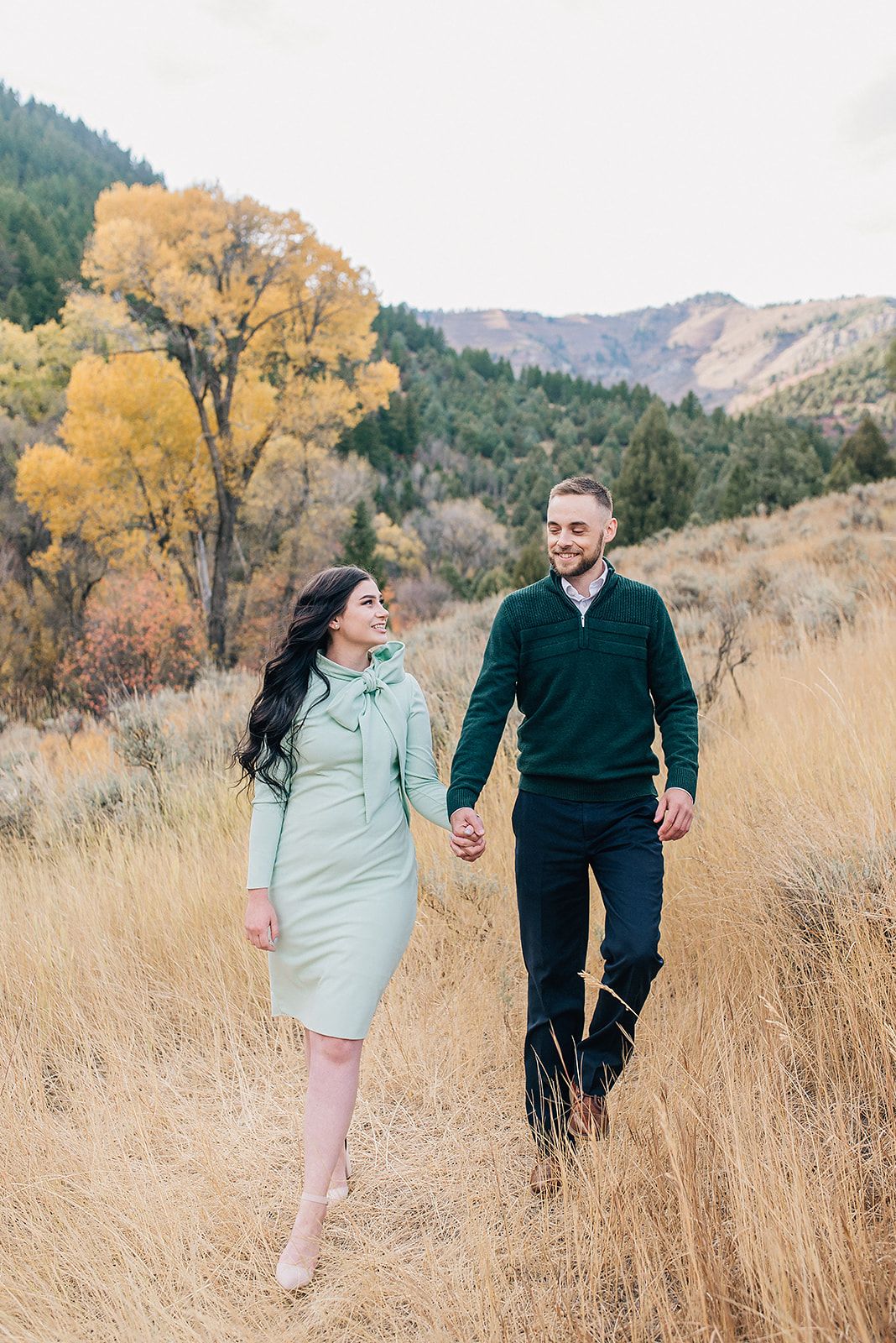 Couple holding hands walking through golden autumn field with colorful mountain foliage backdrop.