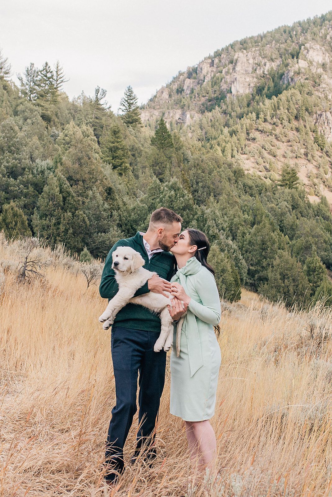 Couple kissing while holding a white puppy in a golden field with a forested mountain backdrop.