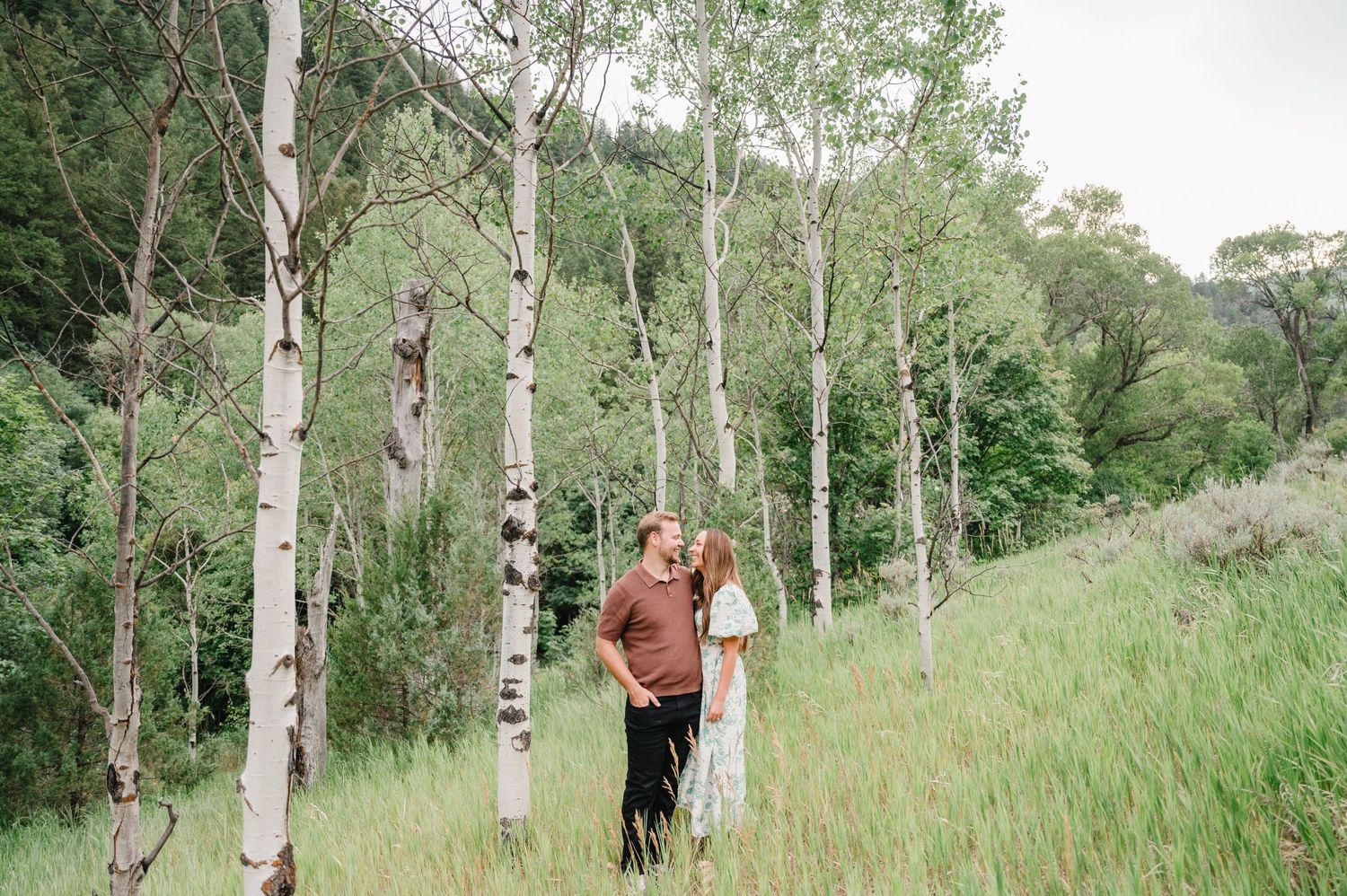 A couple embraces among white birch trees in a lush green mountain setting.
