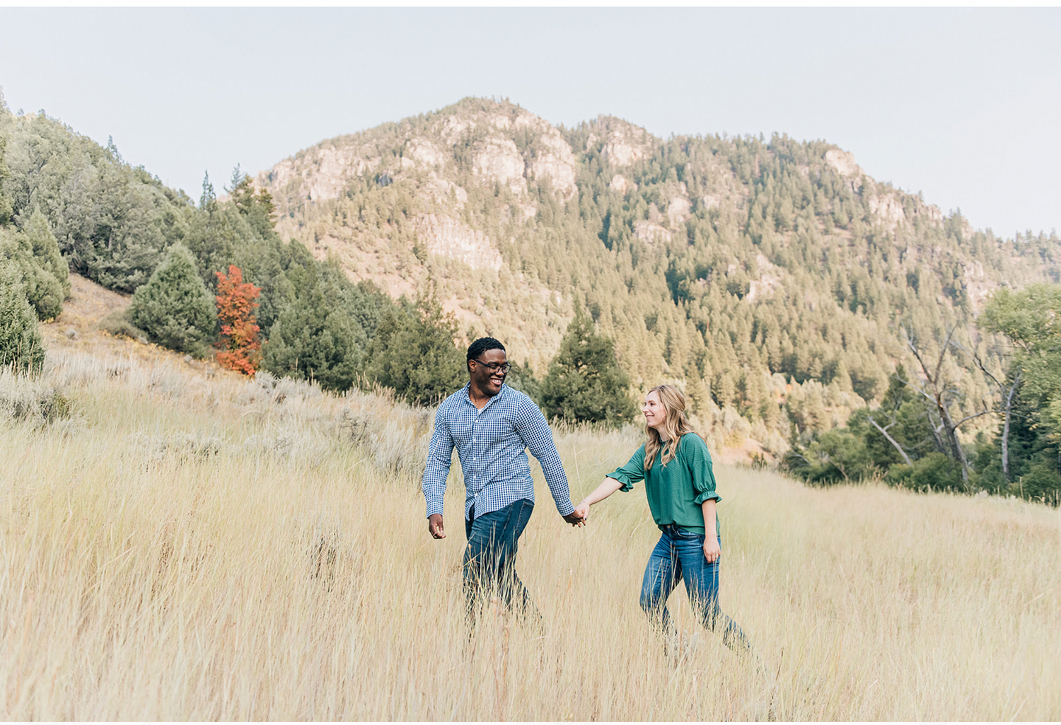A couple walks hand in hand through golden grassland with rugged mountain peaks and evergreen trees in background.