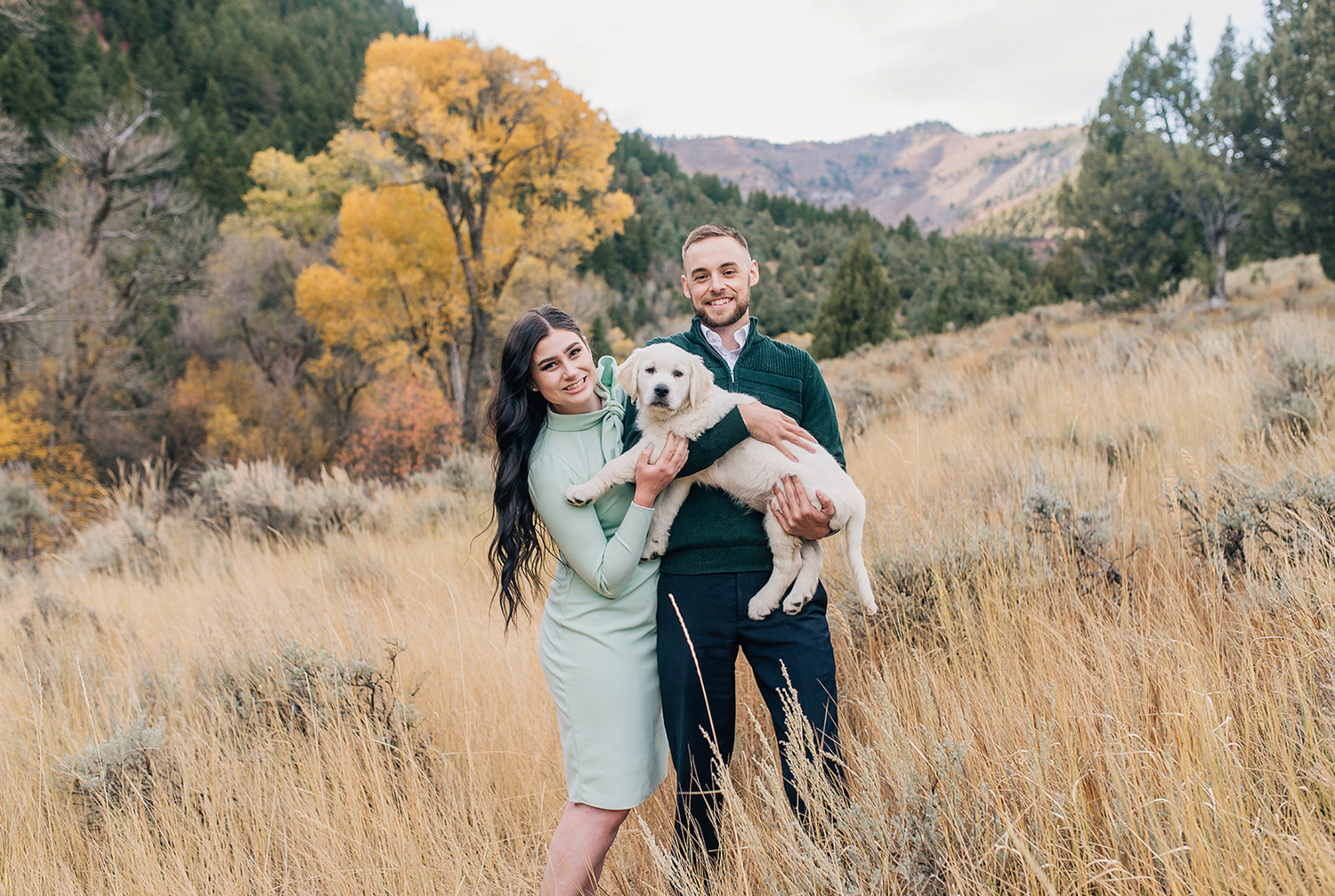 A couple holding their small white dog while standing in autumn meadow with mountain scenery.