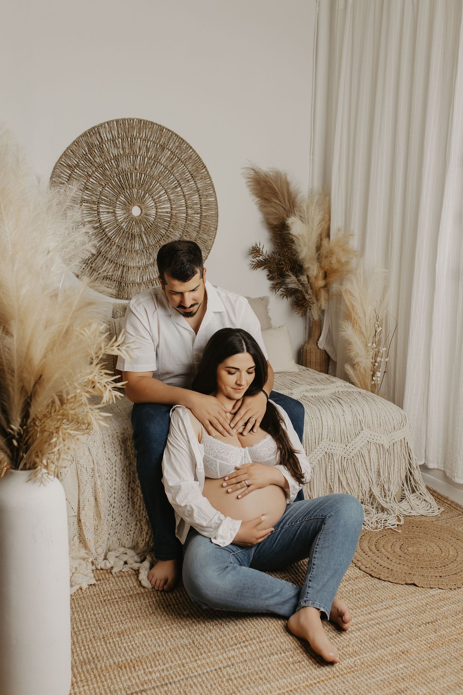 Couple in boho bedroom, pregnant woman sitting on floor cradled by partner, both touching baby bump.