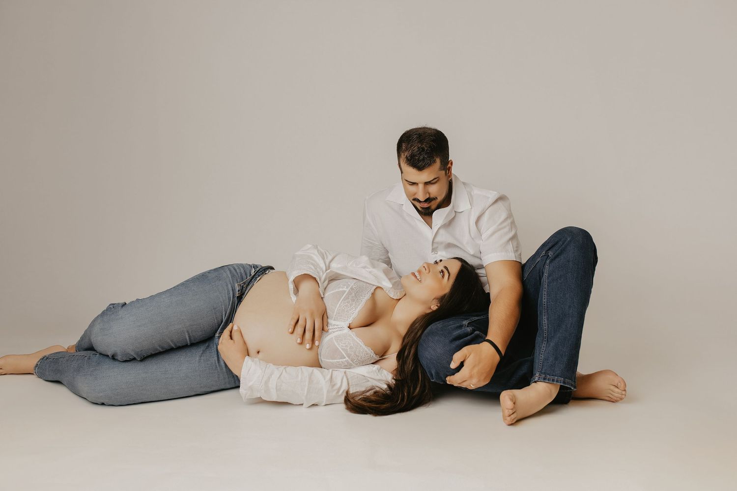 Pregnant woman lying on floor with head on partner's lap during intimate maternity photo session in studio.