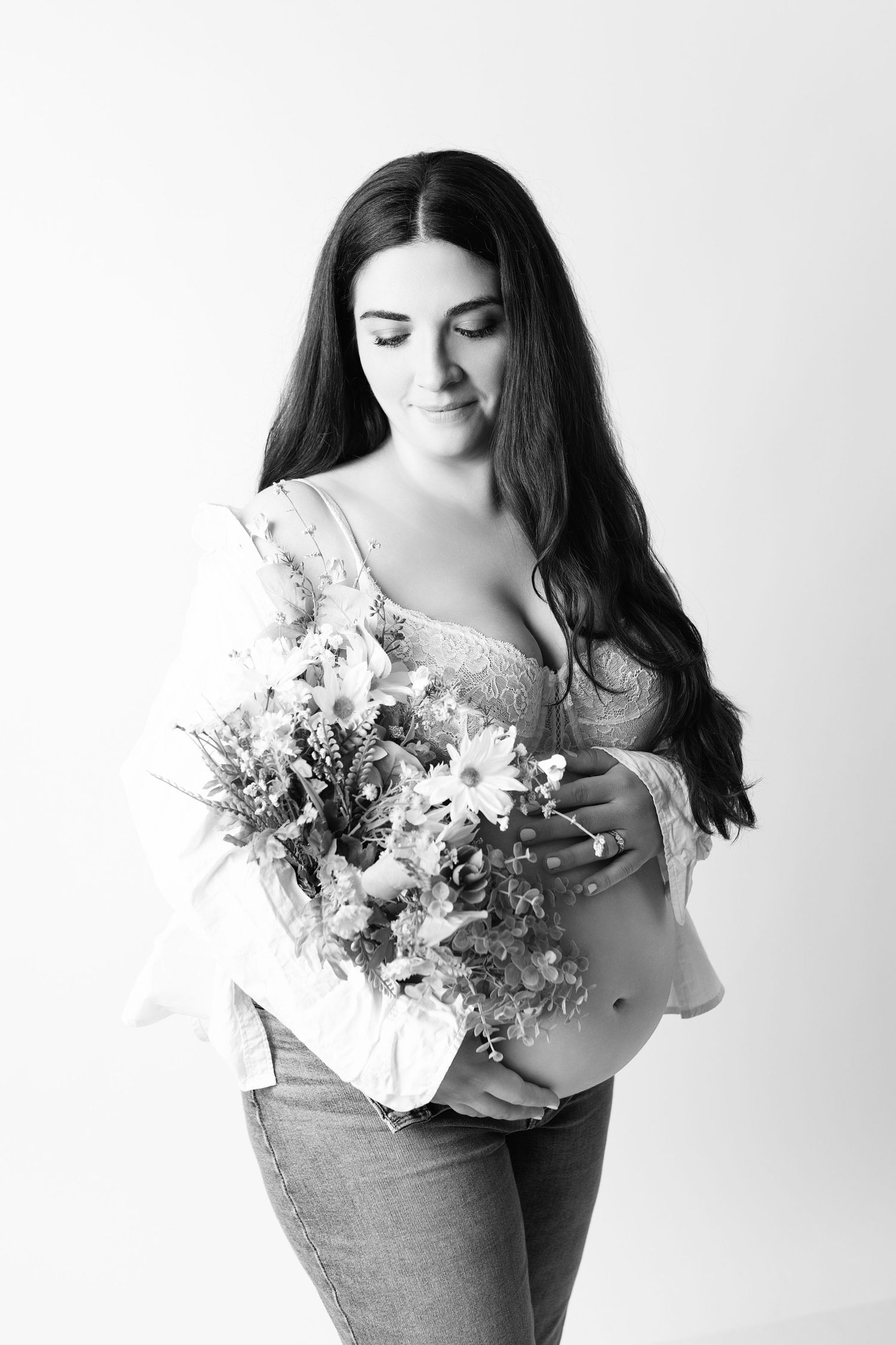 Black and white photo of pregnant woman holding wildflowers, wearing jeans and open shirt, looking down at belly.