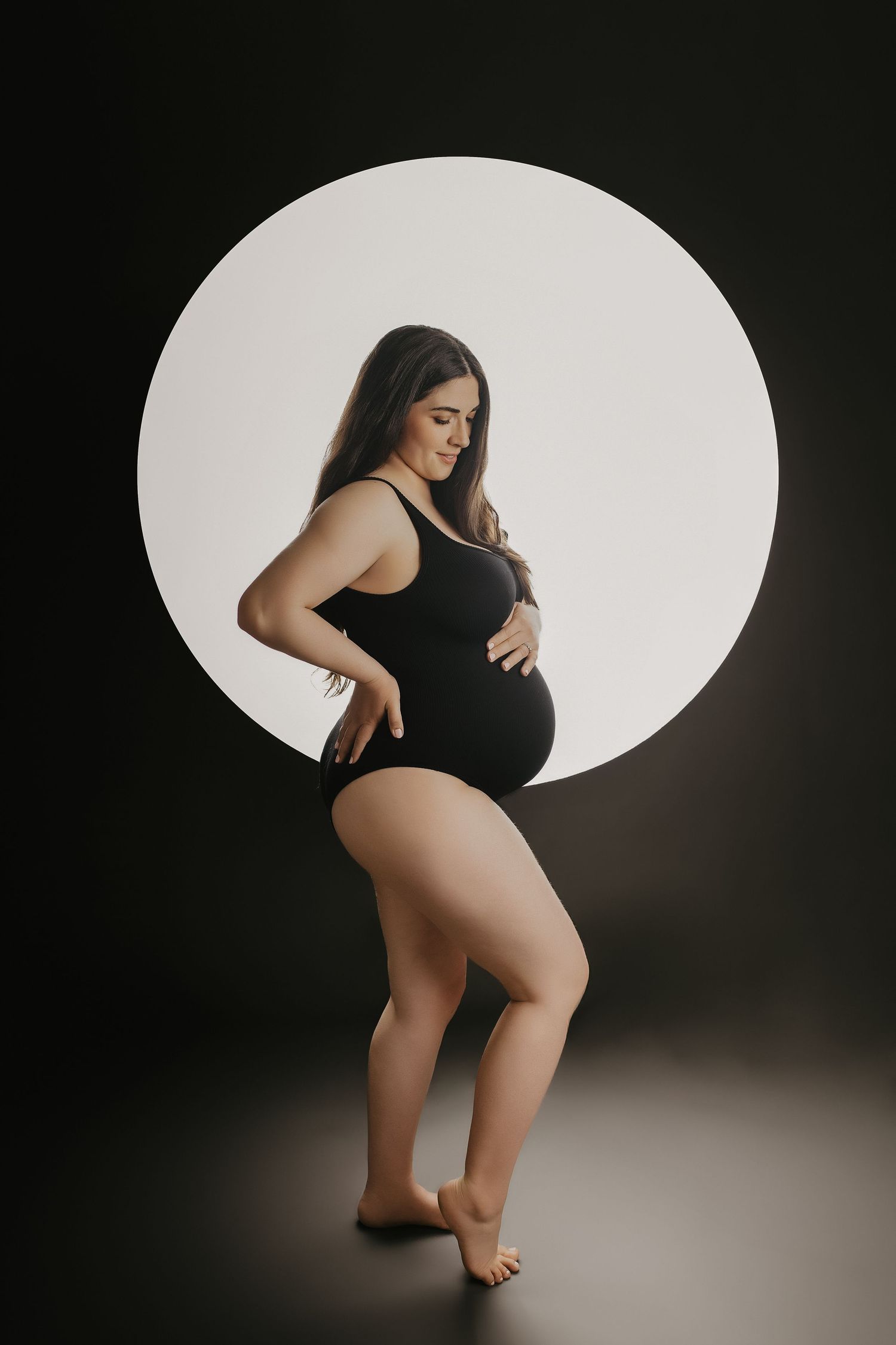 Pregnant woman in black bodysuit posing in front of circular light on dark background.