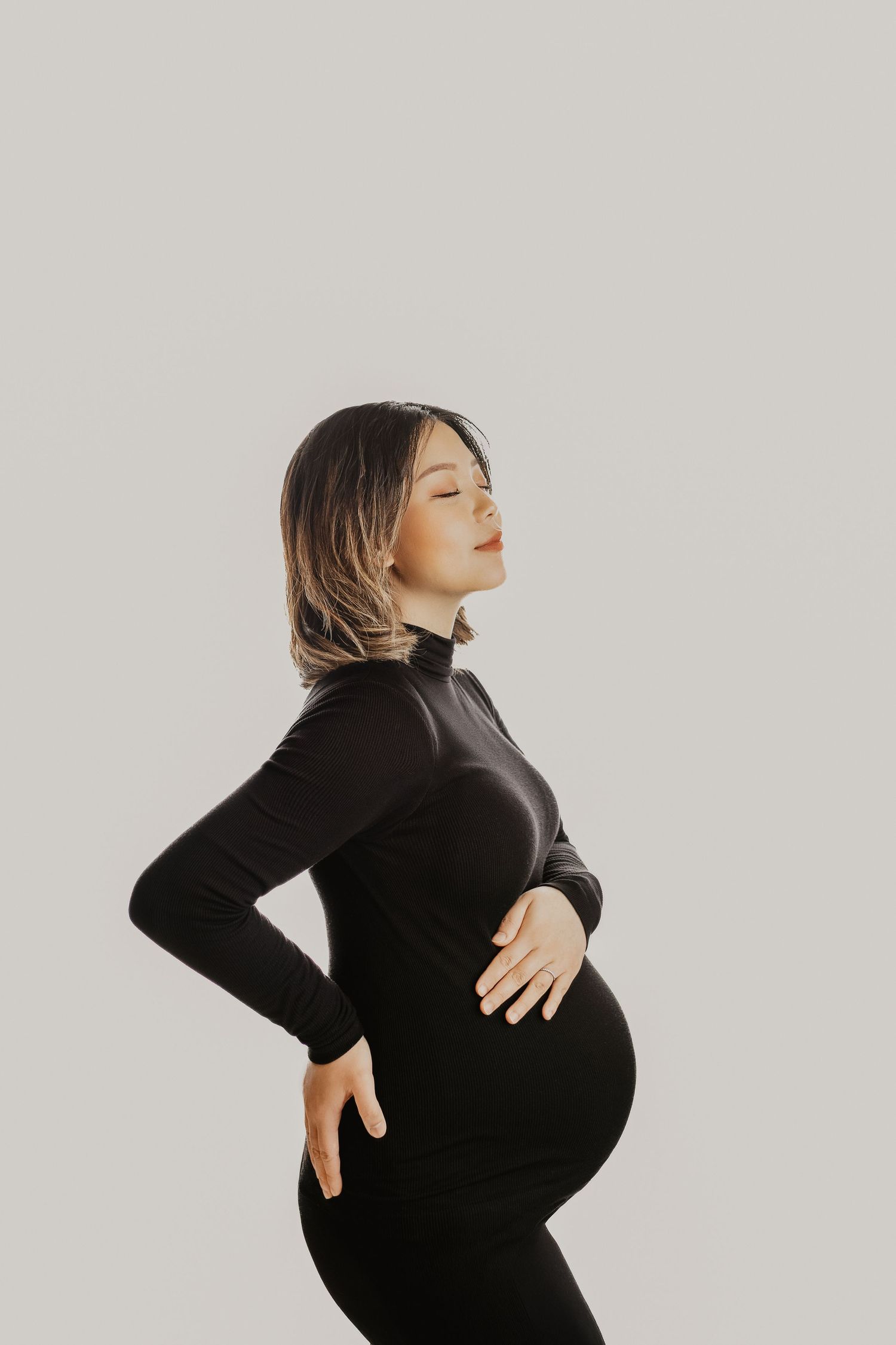 Pregnant woman in black turtleneck outfit holding her baby bump with eyes closed on light background.