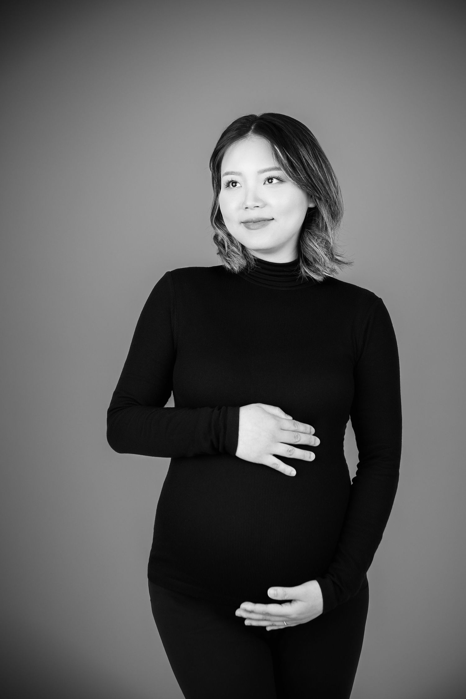 Black and white portrait of a smiling pregnant woman in a black turtleneck, cradling her baby bump.
