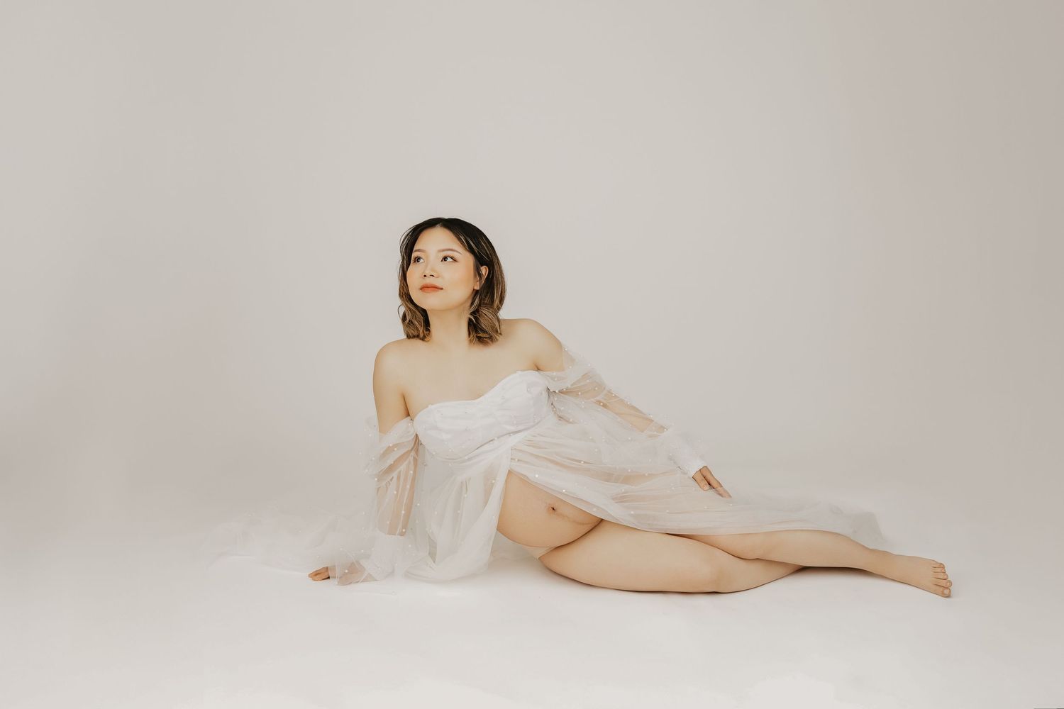 Pregnant woman sitting on floor draped in sheer white fabric against a light gray background in a maternity photo.